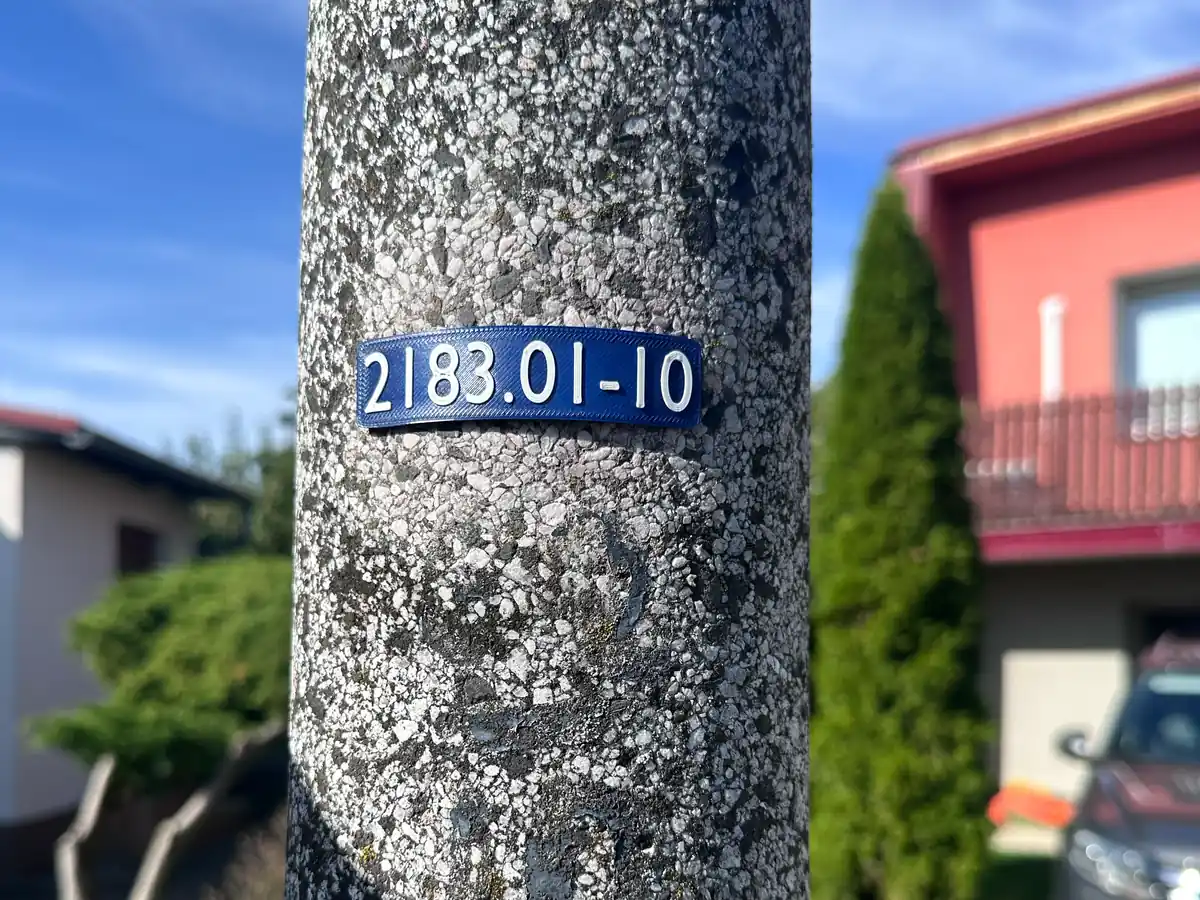 Tmavě modrý štítek s bílým číslem 2183.01-10 nalepený na betonovém stožáru veřejného osvětlení, v pozadí rodinné domy a modrá obloha.