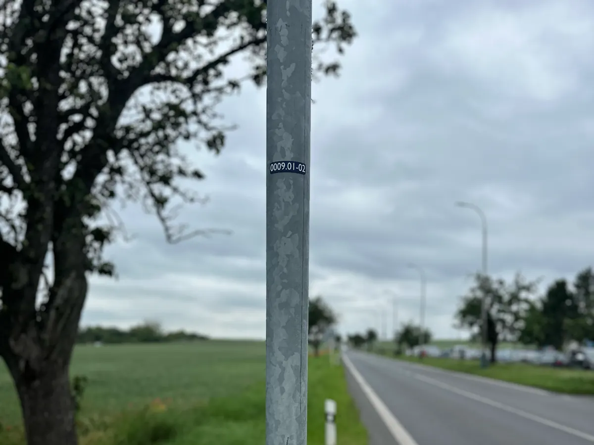 Stožár veřejného osvětlení u silnice mimo obec se štítkem 0009.01-02, číslo je čitelné i od vozovky.