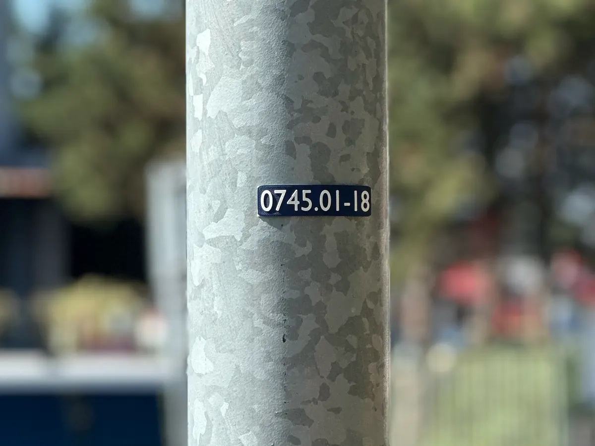 Štítek 0745.01-18 na pozinkovaném stožáru veřejného osvětlení, za ním rozostřená ulice.