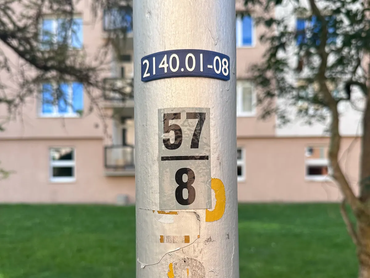 Štítek 2140.01-08 na stožáru na sídlišti, pod ním zbytek původní papírové cedulky s číslem 57/8.