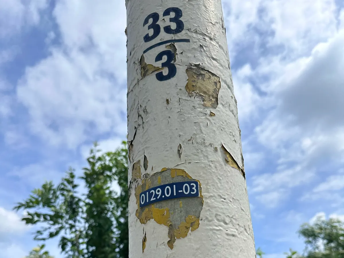Štítek 0129.01-03 nalepený na starém stožáru s oprýskaným bílým a žlutým nátěrem, nad ním původní ručně malované číslo 33/3.