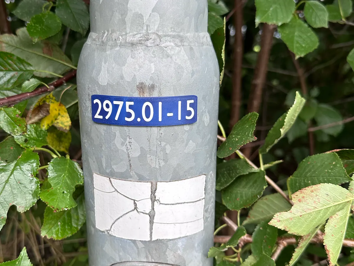 Štítek 2975.01-15 na pozinkovaném stožáru zarostlém keřem, kolem mokré listy.